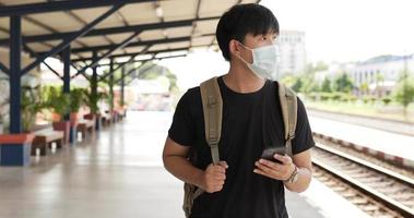 vista frontal de un joven viajero asiático charlando en un smartphone mientras camina en la estación de tren. amigo de encuentro masculino en la estación de tren. concepto de vacaciones, viajes y pasatiempos.