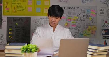 Portrait of Serious asian businessman using laptop and looking document while sitting workplace desk at home office. Young man working at home. Working from home overload at night in new normal.