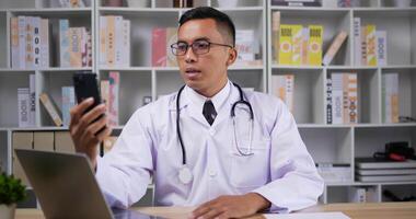 retrato de un médico asiático profesional con bata médica blanca. gafas de hombre haciendo llamadas de conferencia en el teléfono inteligente. consultando al paciente a distancia en línea por cámara web. concepto de telemedicina. video