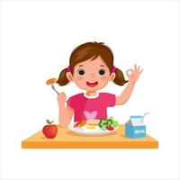 linda niñita desayunando con pan, huevo, brócoli y sosteniendo salchichas con un tenedor que muestra un buen signo vector