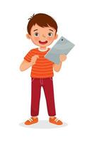 Happy little boy holding and using digital tablet touching screen browsing internet, doing homework, and playing games. Kids and electronic gadget devices concept for children vector