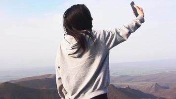 Zoom out view young attractive brunette takes selfie smart phone with scenic landscape mountains background.