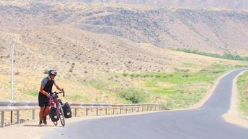 Statische Ansicht Kaukasischer männlicher Fahrradsolo schiebt rotes Fahrrad bergauf auf Asphalt bei extremer Hitze im Freien in malerischen Bergen. Fahrradtouren-Abenteuer-Lifestyle-Konzept video