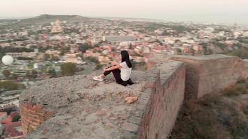 Luftkreis um attraktive weibliche Person, die auf der Burgmauer von Narikala sitzt und den Blick auf die Stadt Tiflis genießt. Georgia