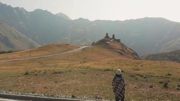 vue arrière d'une touriste debout à distance et regarde vers le point de repère de kazbegi - église de la trinité de gergeti video