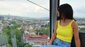visão estática jovem turista caucasiana olha pela janela para a cidade velha em teleférico, tbilisi, geórgia