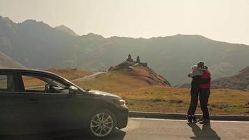 Young caucasian couple stands and hugs by the car in scenic location. Kazbegi road trip vacation concept