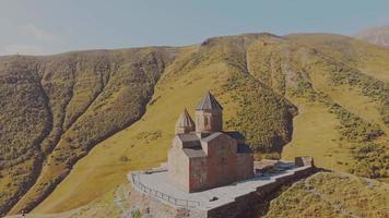 círculo aéreo de câmera lenta ao redor da igreja da trindade kazbegi na colina com fundo de montanhas do cáucaso. locais históricos e conceito de cultura georgiana video