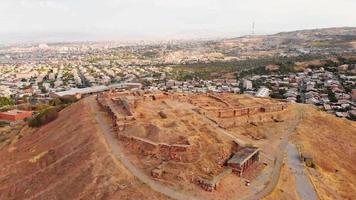 Aerial panning view triangle shape Erebuni fortress ruins on hilltop with Yerevan city panorama background . Old urartian fortified city . Important historical site in Armenia video