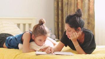 Young mom or babysitter lay on bed and help boy kid with homework reading school notebook sitting on bed in bedroom. Caring mother explain lesson to preschooler child