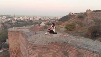 Aerial Side view in motion around female woman sitting on high viewpoint with beautiful background panorama of Tbilisi city. Georgia