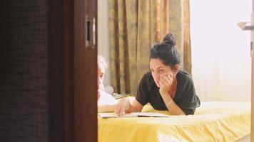 la joven madre o la niñera se acostaron en la cama y ayudaron a un niño con la tarea leyendo el cuaderno de la escuela sentado en la cama en el dormitorio. madre cariñosa explica la lección al niño en edad preescolar video