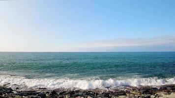 statische weergave real-time golven crashen over rotsen op het strand met horizon en heldere lege blauwe hemelachtergrond video