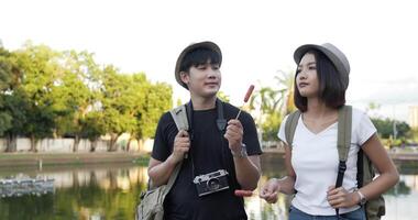 vista lateral de una pareja feliz asiática con sombrero comiendo salchichas mientras camina por el parque. alegre joven y mujer comiendo un apetitoso. concepto de vacaciones y estilo de vida. video