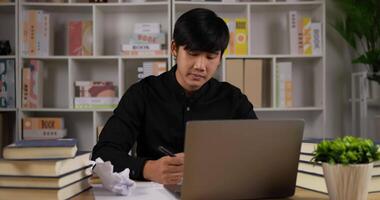 porträt eines erschöpften asiatischen geschäftsmannes, der schreibt und hände reißt papier, während er am arbeitstisch im heimbüro sitzt. schlechtes arbeiten erfolglos, keine ahnung, scheitern konzept. video