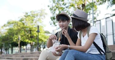 Side view of Happy Couple Asian with hat eating sausages while sitting on stairs at the park. Cheerful Young man and woman eating an appetizing. Vacation and lifestyle concept. video
