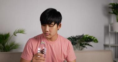 Portrait of Tired Asian man sitting on a couch drink chilly fresh water from a bottle. Health care concept. video