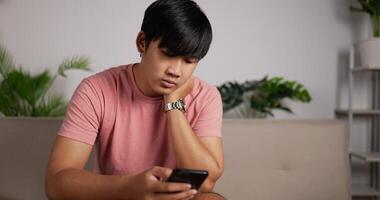 retrato de homem asiático entediado usando smartphone sentado no sofá na sala de estar em casa. homem cansado olhando o usuário de internet do telefone móvel rola através de aplicativos de smartphone. conceito de mídia social video