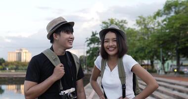 vista lateral de la feliz pareja de viajeros asiáticos con sombrero sonriendo y mirando la cámara en el parque. alegre joven blogger hombre y mujer saludando con cámara en el parque. concepto de hobby y estilo de vida. video