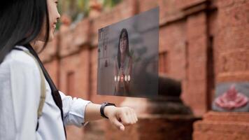 Portrait side view of Asian woman chatting on video call on smartwatch with friend while standing at ancient temple. Projecting visible AR screen and talking to friend online. Futuristic concept.