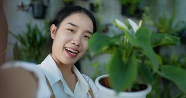 retrato de una joven y feliz jardinera asiática que vende en línea en las redes sociales y mira la cámara en el jardín. selfie de mujer con teléfono móvil. vegetación casera, venta en línea y afición. video