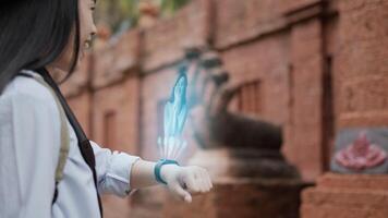 Portrait side view of Asian woman pressing buttons on smart watch while standing at ancient temple. Projecting visible AR screen and chatting on video call. Futuristic and technological concept