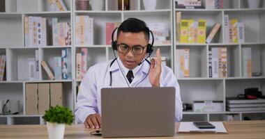 retrato de un médico asiático profesional con bata blanca y auriculares haciendo una llamada de conferencia en una computadora portátil en la habitación. consultando al paciente a distancia en línea por cámara web. concepto de telemedicina. video