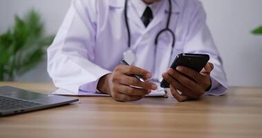 Close up of Professional male doctor in white medical coat using smartphone, web surfing information online or giving distant consultation to patient, writing notes on clipboard paper in the clinic. video