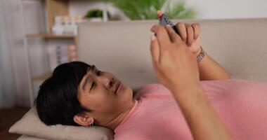 un jeune homme asiatique heureux qui regarde un film sur son smartphone se détend sur un canapé à la maison. un homme souriant utilisant des applications de médias sociaux mobiles regarde un téléphone portable en profitant de son temps libre avec un appareil technologique. passe-temps et vacances. video