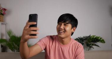 portret van glimlachende aziatische die een person.young man videogesprek belt en praat met smartphone in huis woonkamer. ontspannen man die op mobiele telefoon spreekt. bespreken met vriend familie. video