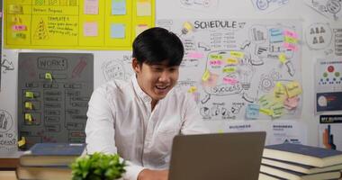 empresário asiático profissional senta-se na mesa do local de trabalho com um laptop trabalhando no escritório. ele recebe ótimas notícias e fica encantado. masculino sorrindo e braços cruzados. arranque e conceito de empresário. video