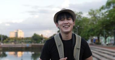 Vista lateral do homem feliz viajante asiático com chapéu sorrindo e olhando para a câmera no parque. homem jovem blogueiro alegre rindo com a câmera no parque. conceito de hobby e estilo de vida.