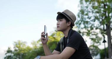 retrato de vista lateral de homem feliz jovem viajante asiático com chapéu falando no celular enquanto está sentado na escada no parque. homem sorridente falando no celular no parque. conceito de hobby e estilo de vida.