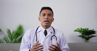 Asian doctor talking to patient making video call on laptop while sitting on sofa. Professional physician speaking looking at camera by webcam in web chat consulting client online.Telemedicine concept
