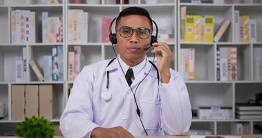 portrait d'un médecin de sexe masculin asiatique professionnel en blouse médicale blanche. casque d'homme faisant une conférence téléphonique et regardant la caméra. consultation patient à distance en ligne par webcam. notion de télémédecine. video