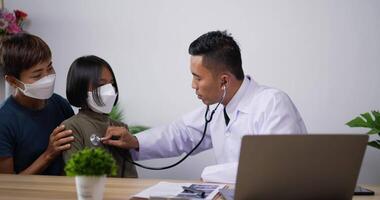 Médico pediatra asiático profesional que usa estetoscopio escucha el corazón de una niña pequeña con mascarilla facial y madre en la clínica. médico varón examinando a un niño. concepto médico y de salud. video