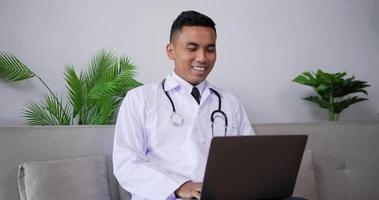 Asian doctor talking to patient making video call on laptop while sitting on sofa. Telemedicine male physician speaking looking at screen computer by webcam in web chat consulting client online.