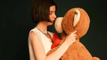A young brunette girl, with short hair, in a white blouse is holding a large red teddy bear in her hands.