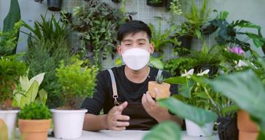 portret van een gelukkige jonge aziatische mannelijke tuinman die op sociale media verkoopt en naar de camera kijkt. man in gezichtsmasker met plantpot. thuisgroen, online winkelen en hobbyconcept. video