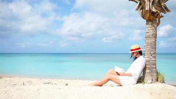 Lesebuch der jungen Frau auf weißem Strand, der unter der Palme sitzt