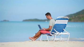 Young man with laptop on tropical caribbean beach. Man sitting on the sunbed with computer and working on the beach video