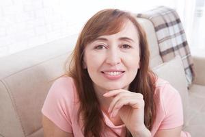 retrato macro de rostro femenino sonriente. atractiva y hermosa mujer de mediana edad sentada en un sofá y relajándose en casa. menopausia. día de la Madre foto