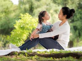 niña asiática sentada con su madre en la tela, relájese y aprenda fuera de la escuela para disfrutar en el parque natural foto
