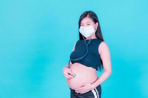 las mujeres asiáticas embarazadas usan una máscara y usan un estetoscopio para tocarse el vientre y escuchar a su bebé por nacer foto