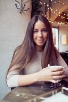 Beautiful pensive happy girl sitting in cafe in Christmas holidays, smiling and dreaming. Brunette woman with long hair drinks cappuccino coffee, latte and looks to camera photo