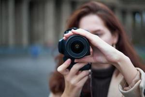 Selective focus on lens. Beautiful stylish fashionable girl holds camera in her hands and takes pictures. Woman photographer with long dark hair in city, urban shoot, vertical. unrecognizable person photo