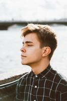 Portrait of young guy, brunette with dark hair, Caucasian. Close - up of a guy, boy, man looking past camera. Outdoor in city by water photo