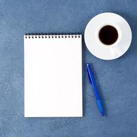 libreta abierta con página blanca limpia, bolígrafo y taza de café sobre una mesa de piedra azul oscuro envejecida, vista superior