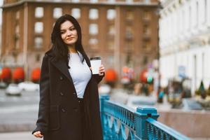 Beautiful serious smart brunette girl holding cup of coffee in hands goes walking down street in center on blue bridge. Charming thoughtful woman with long hair wanders alone, immersed in thoughts photo