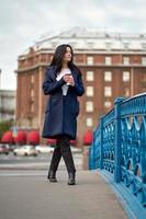 Beautiful serious smart brunette girl holding cup of coffee in hands goes walking down street of St. Petersburg in city center on bridge. Charming thoughtful woman with long dark hair wanders alone photo